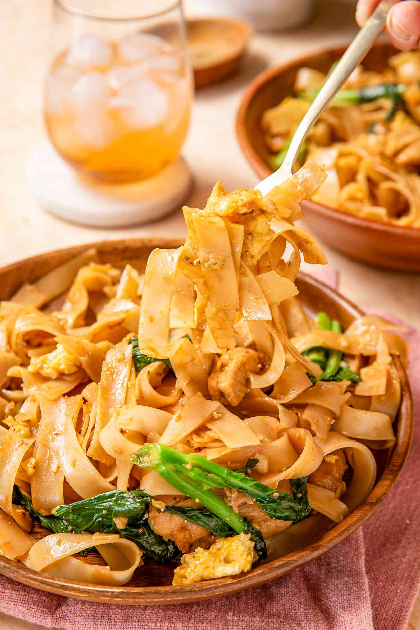 pad see ew in a bowl with a fork lifting some from the plate at a table setting with another bowl of pad see ew and an iced drink in the background