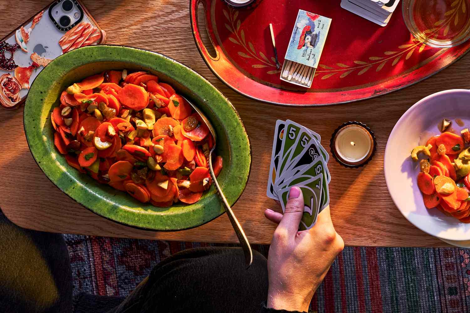 A coffee table with a bowl of carrot salad, cell phone, candles and matchbook, uno cards