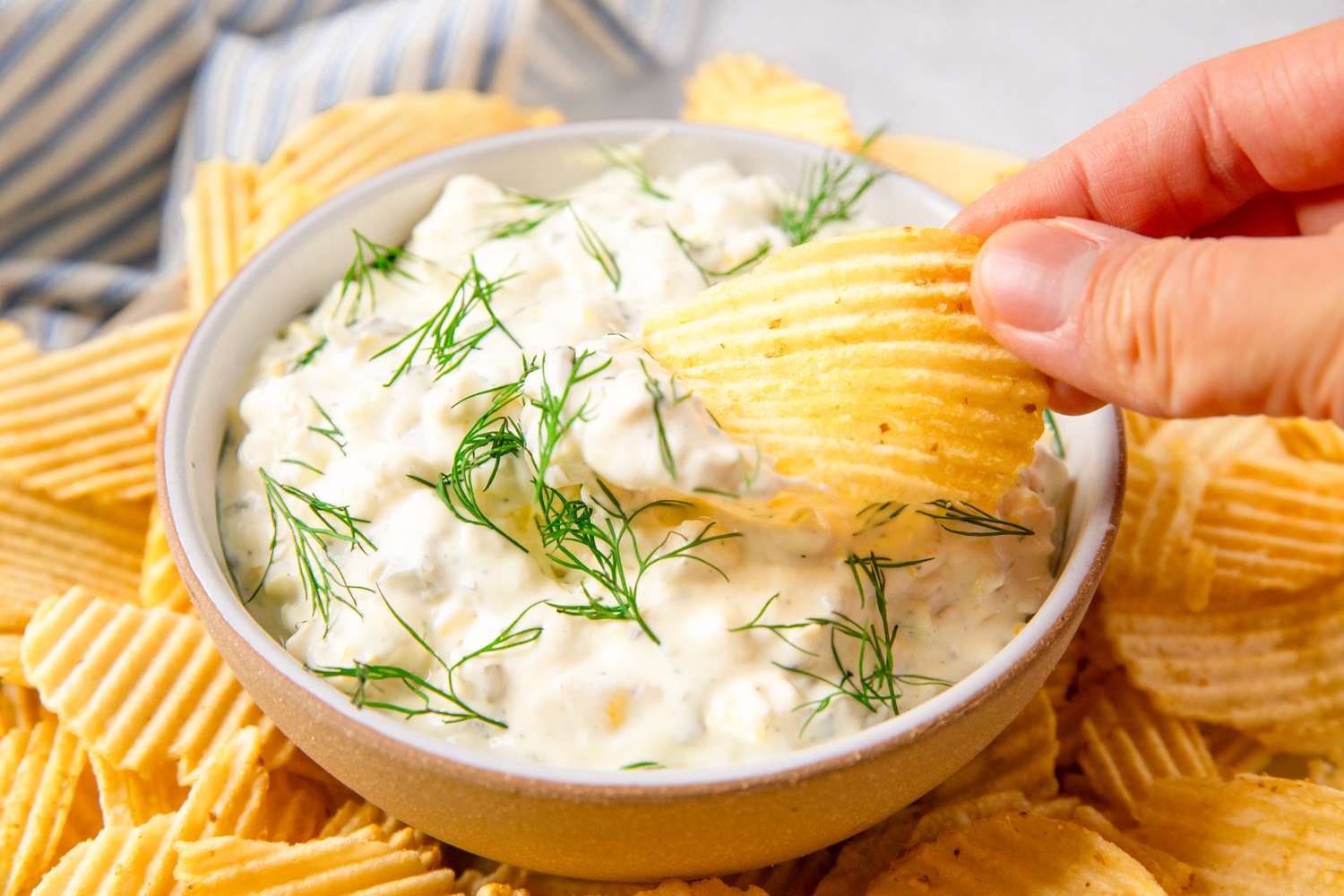 A bowl of creamy dip garnished with fresh herbs, surrounded by crinkled chips, with a hand dipping one of the chips