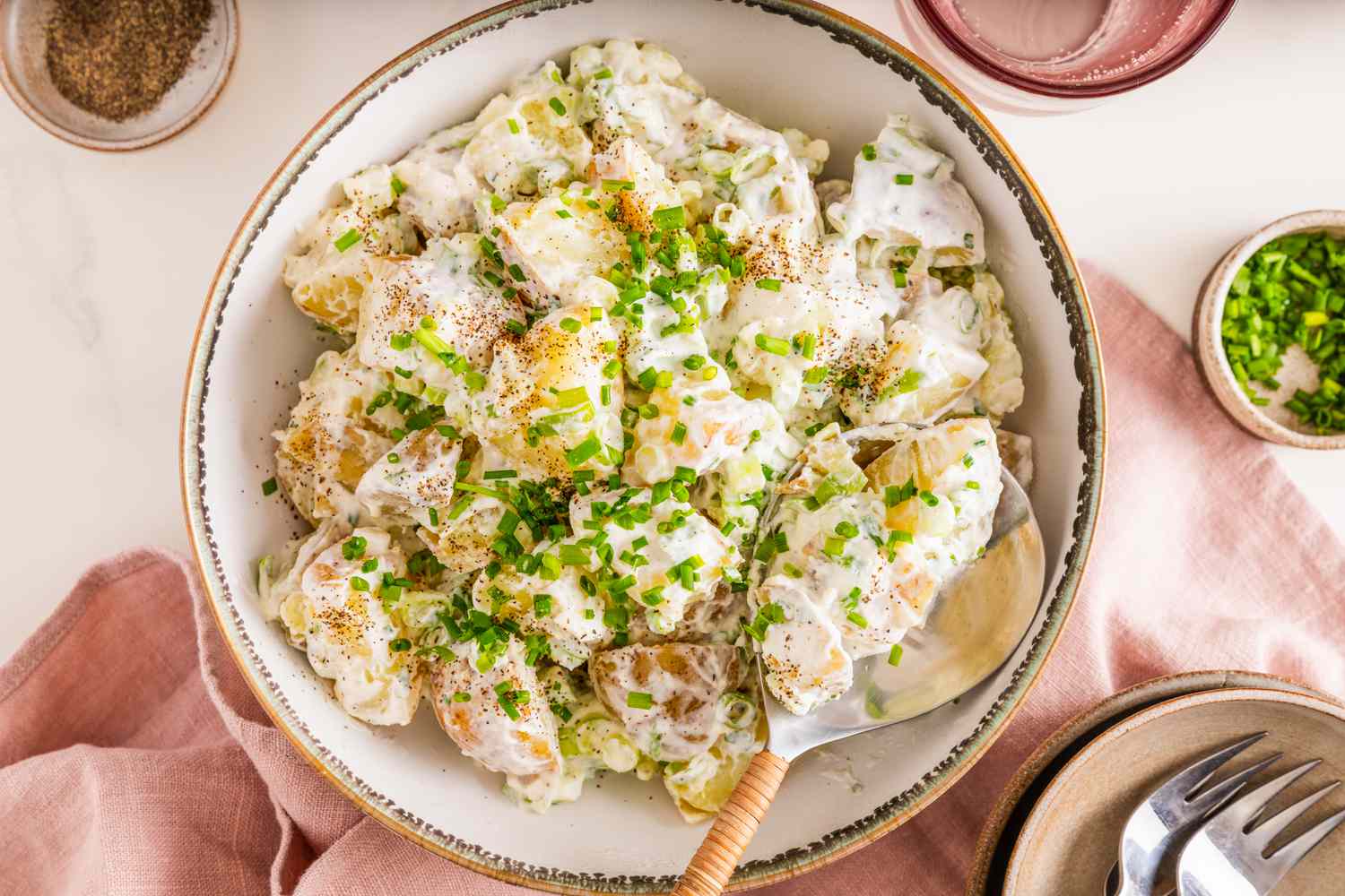 Bowl of Sour Cream and Onion Potato Salad With a Spoon, and in the Surroundings, a Bowl of Seasoning, a Glass of Water, a Bowl of Chives, Stack of Plates With Utensils, and a Blush Table Napkin