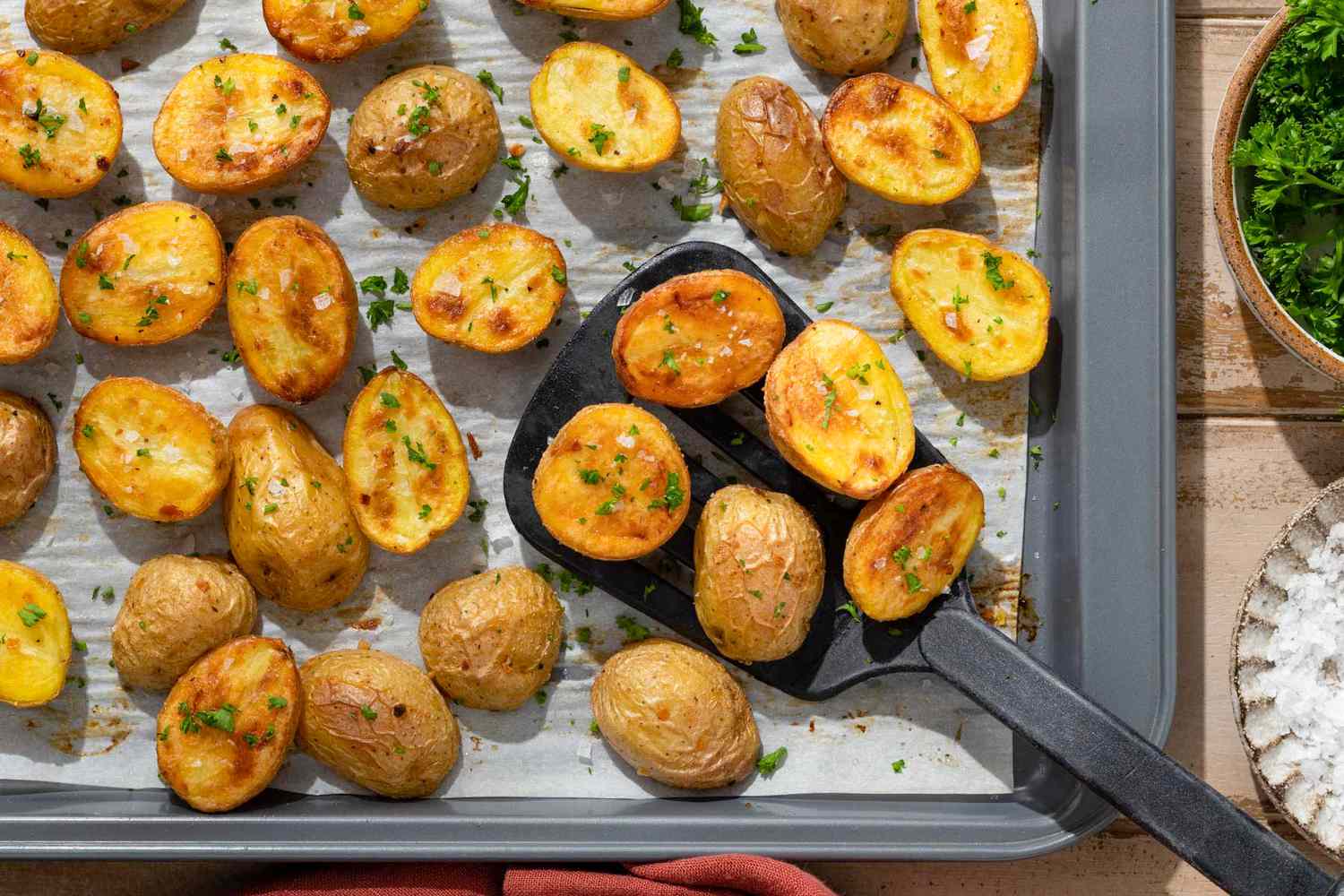 Roasted halved potatoes on a baking sheet, sprinkled with flakey salt and parsley, getting scooped up with a spatula