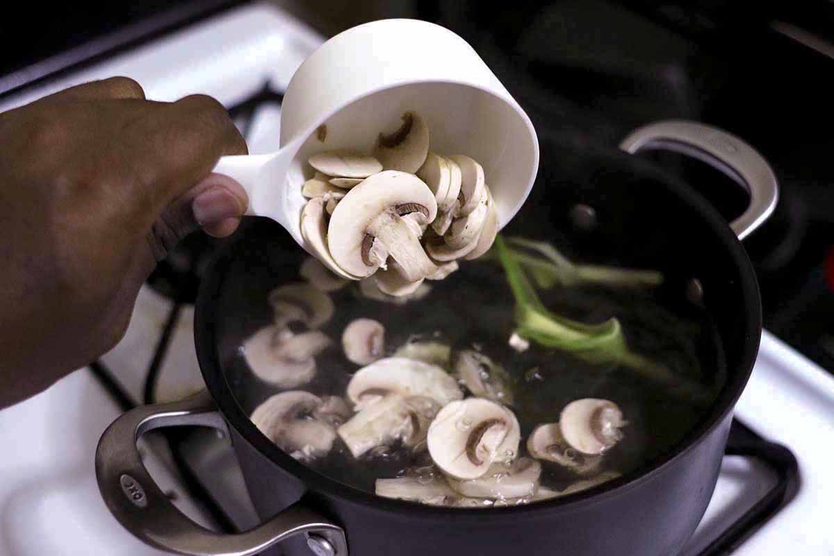 Sliced mushrooms are being added to a black pot on the stove for tom yum soup.