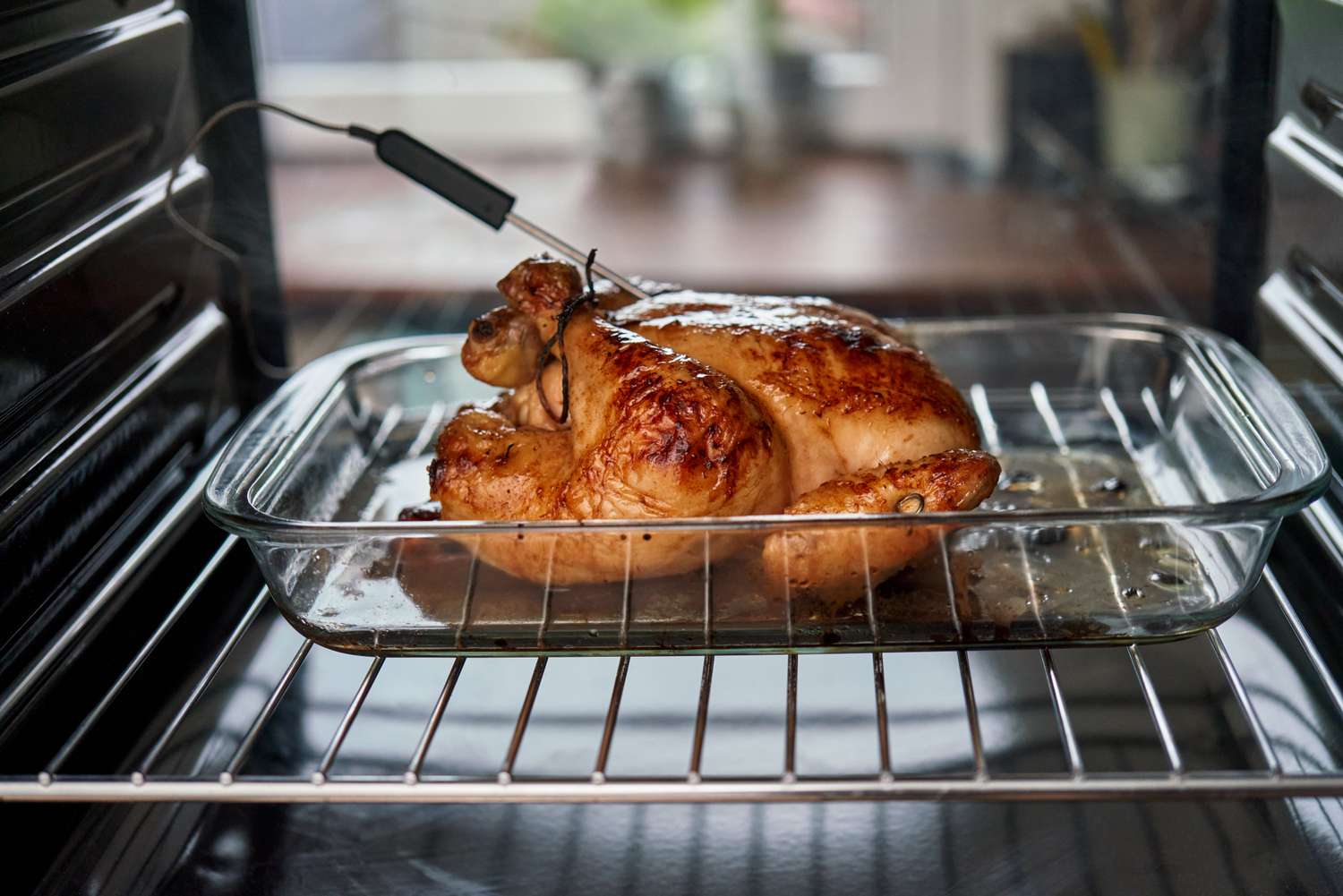 A roasted chicken is in a glass baking dish inside an oven with a meat thermometer inserted into it