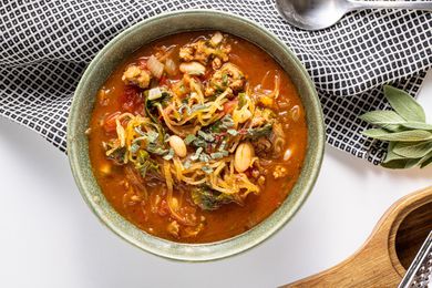 Spaghetti Squash Soup with Italian Sausage in a Bowl Next to a Kitchen Towel, a Spoon, Fresh Sage Leaves, and a Wooden Holder with a Grater