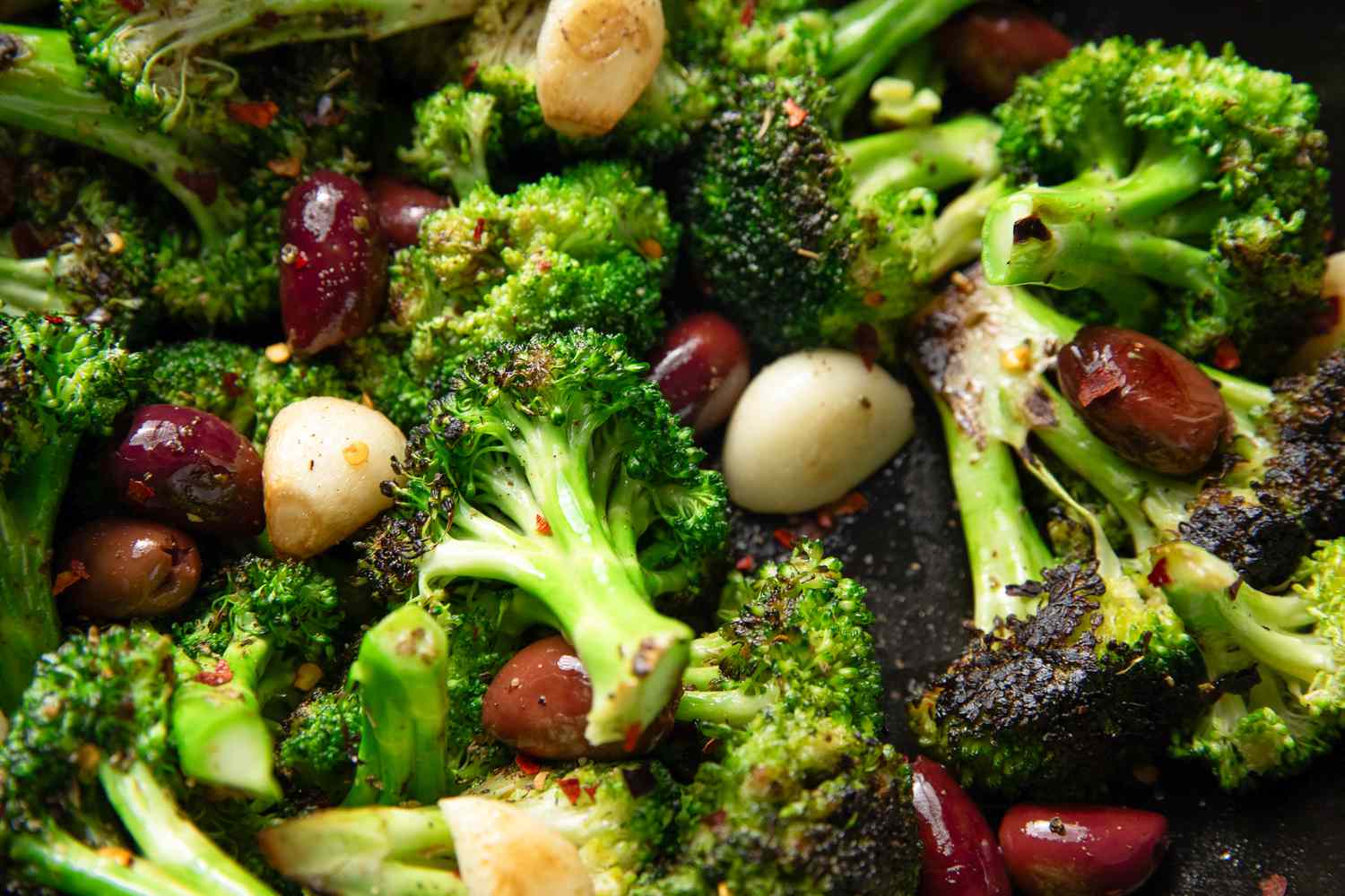 Closeup view of a cast iron skillet of seared broccoli with spices, garlic cloves and olives