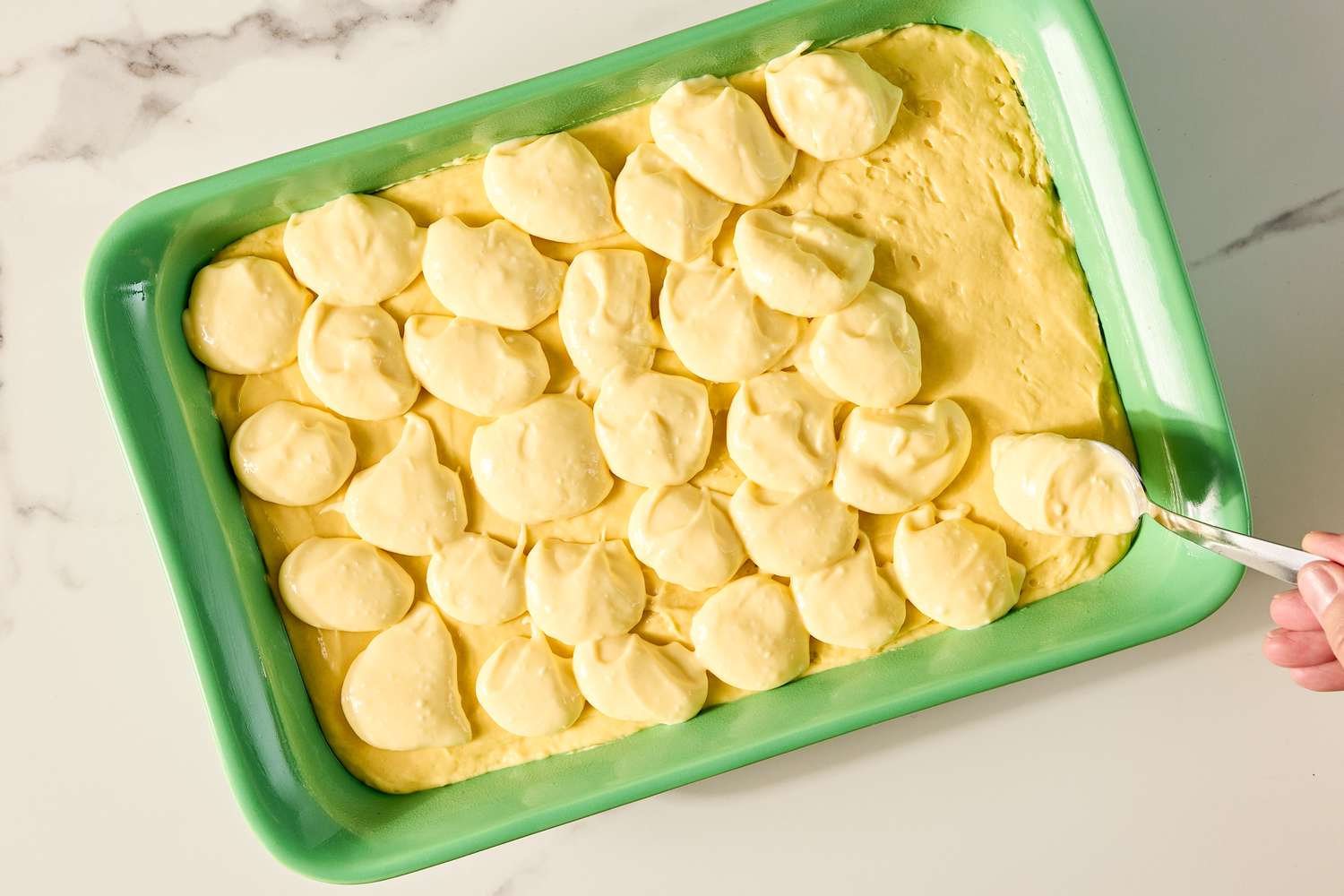 A hand spreading cake batter for the ugly cake recipe in a green baking dish with dollops of filling