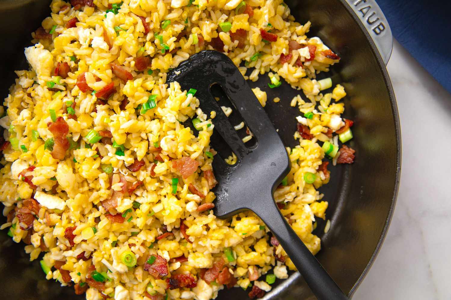 A pan of fried rice with chopped green onions and bits of meat, being stirred with a black spatula