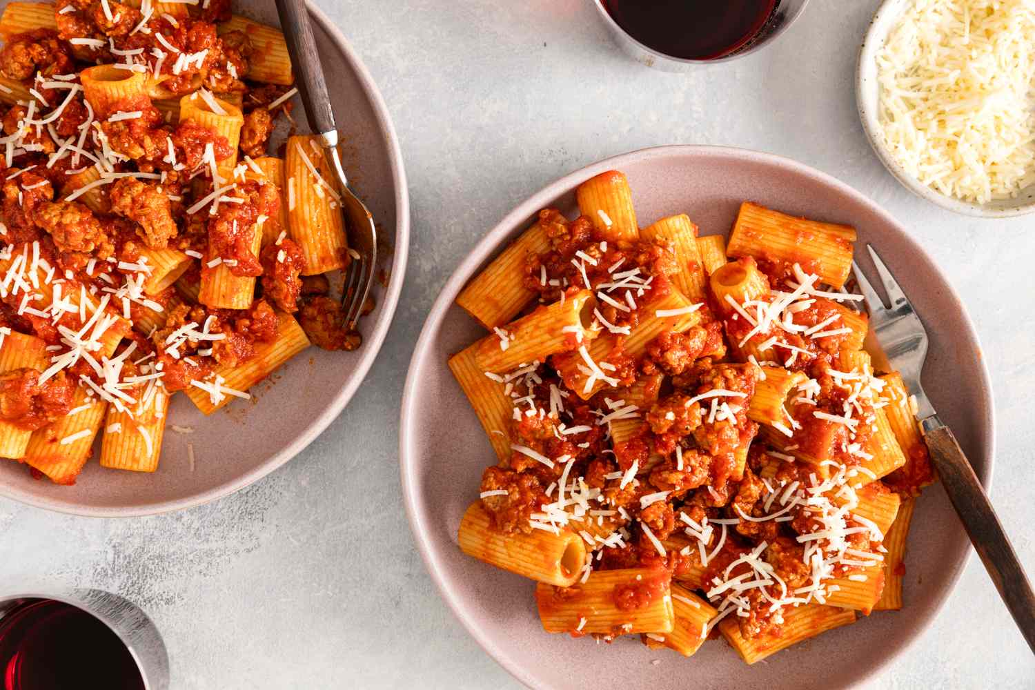 Plates of rigatoni pasta with sausage and tomato sauce topped with grated cheese served alongside glasses of red wine