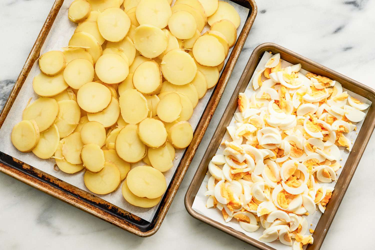 sliced potatoes on a parchment lined sheet-pan, sliced boiled eggs on a smaller parchment lined sheet-pan for Rakott Krumpli (Hungarian Potato Casserole)