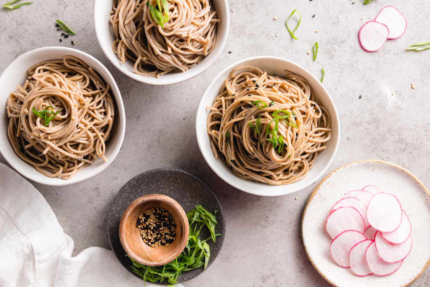 Bowls of Soba Noodles with Sesame Sauce Next to Plate with Spring Onions and Seasoning and Another Plate with Sliced Radish