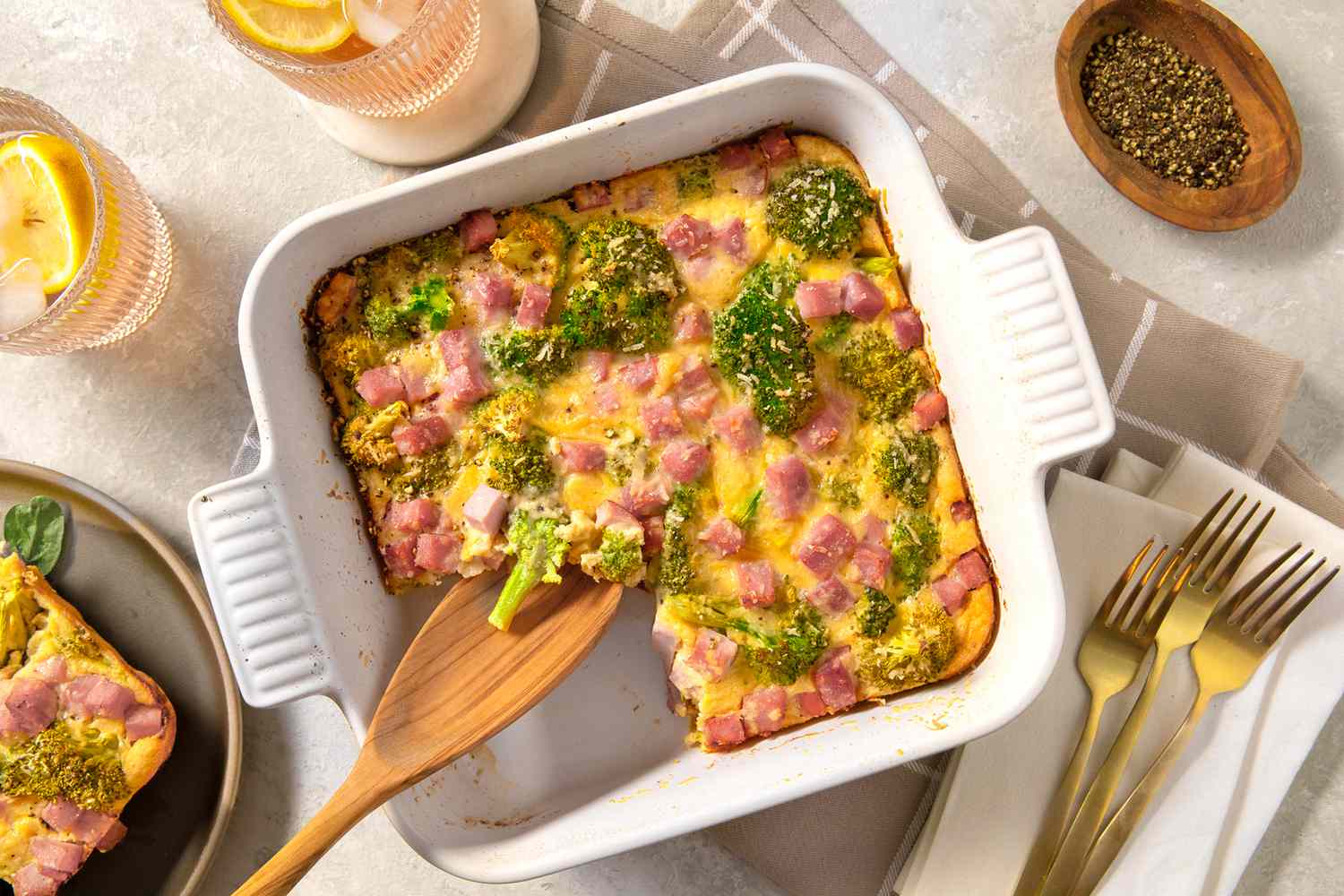 Overhead view of a white casserole dish of broccoli cheese pie with a serving spoon in a table setting with forks, glasses and a bowl of pepper to the side