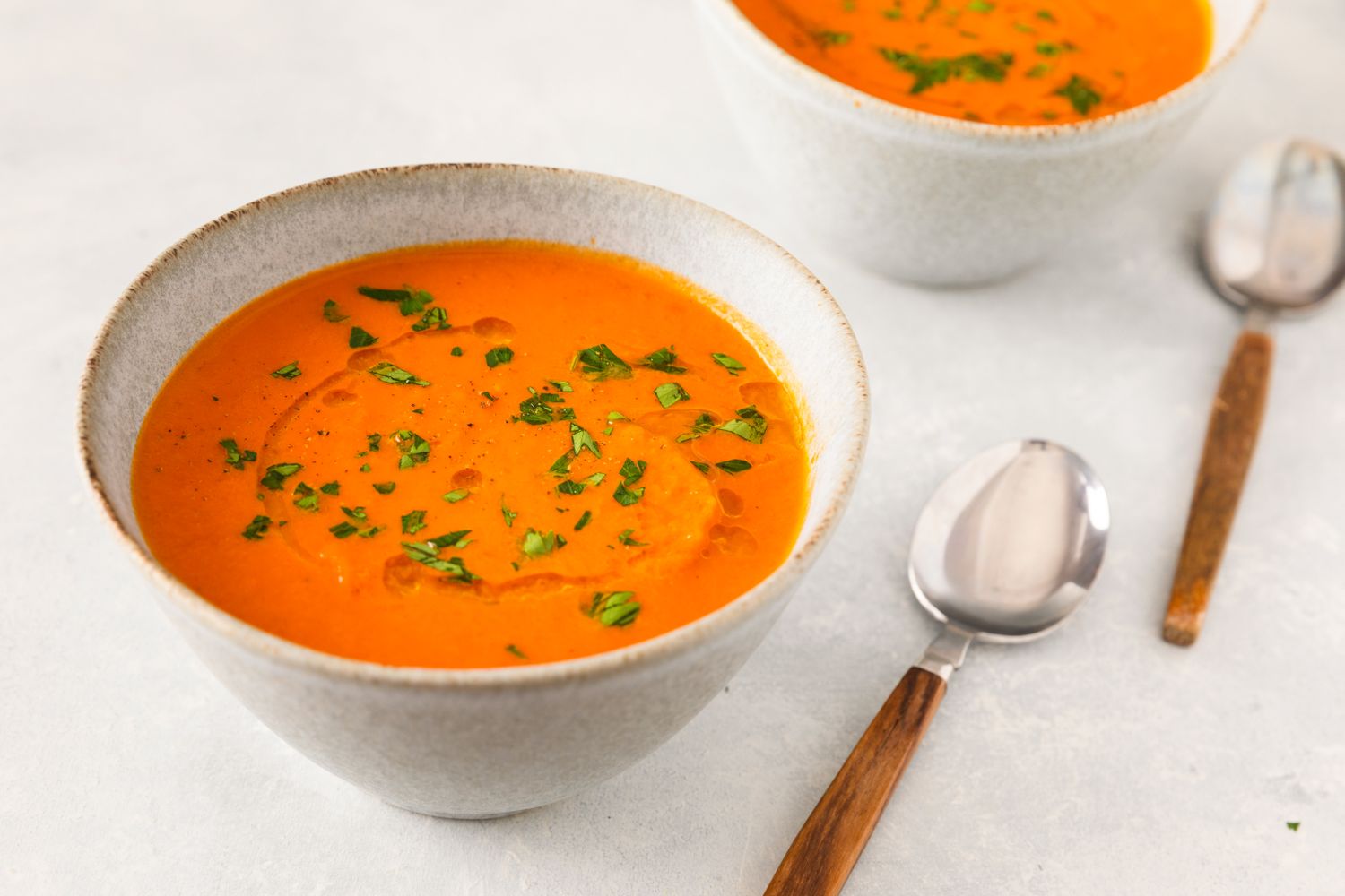 Two bowls of pumpkin tomato soup garnished with herbs served with spoons on a light surface