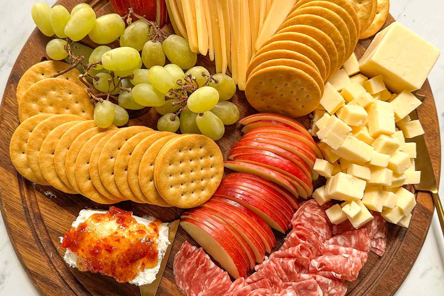 An arranged charcuterie board with crackers cheese grapes apple slices cured meats and a spread with jelly topping on bread