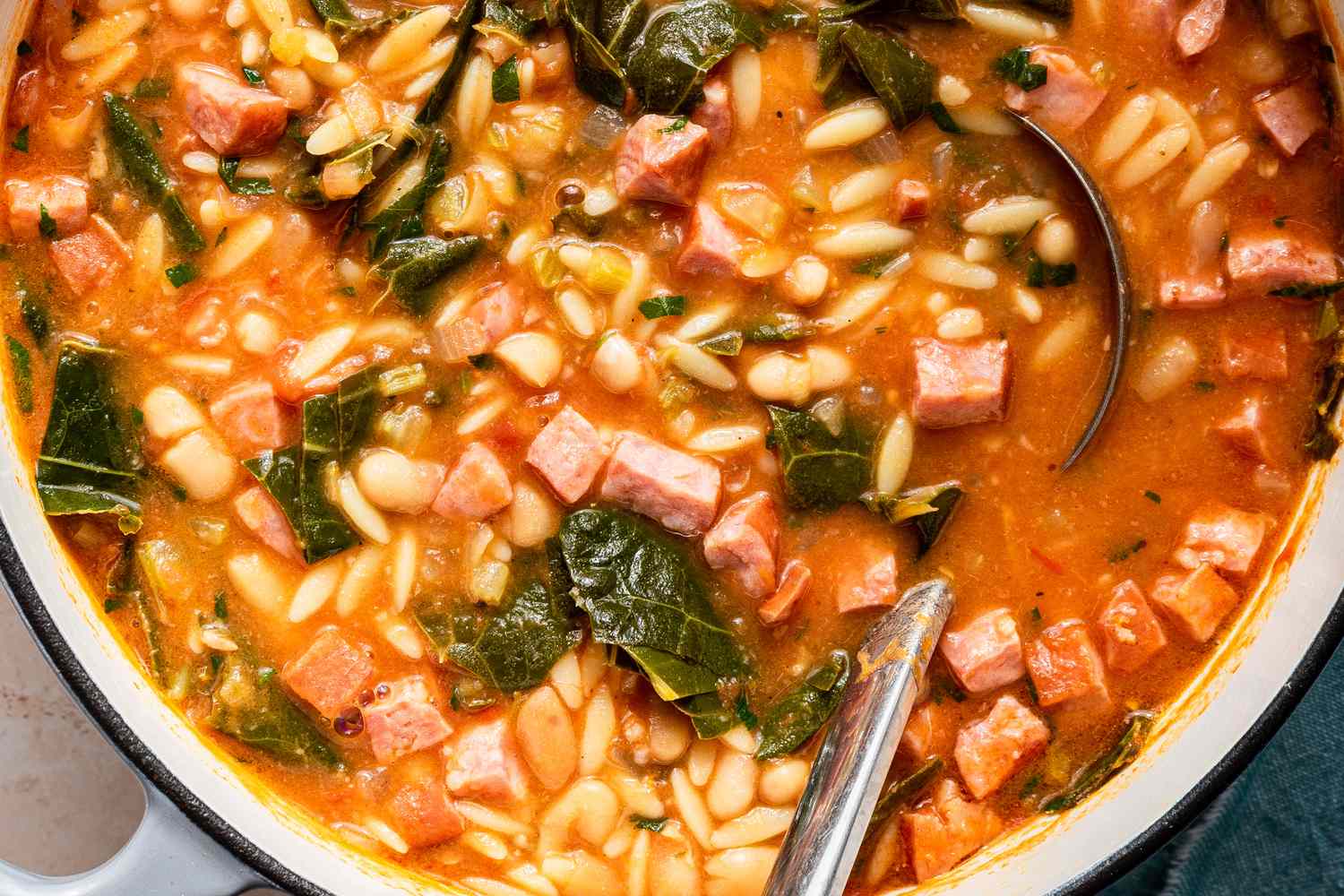 Pot of soup with beans orzo diced ham and greens served with a ladle and spoon
