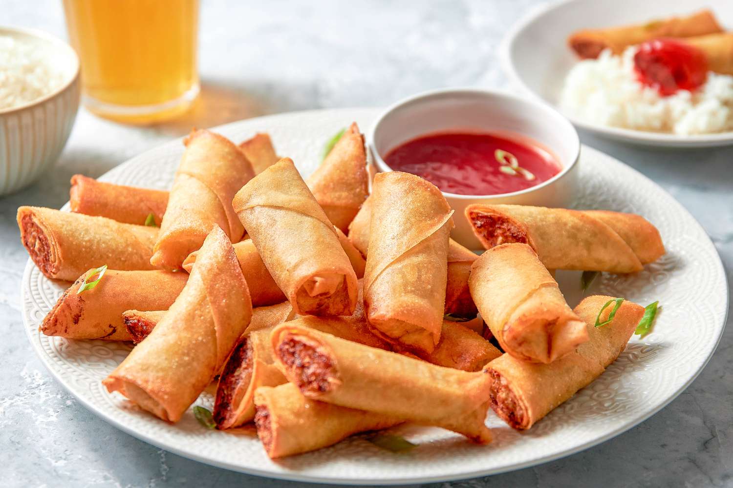 Plate of Lumpia Cut in Half With a Bowl of Sweet and Sour Sauce, and in the Background, a Bowl of Rice, a Glass With a Fizzy Drink, and a Plate With Rice, Lumpia, and Some Sauce