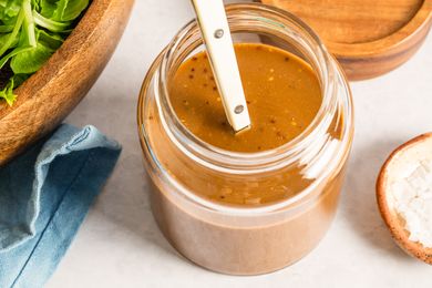 A glass jar of balsamic dressing with a spoon placed next to a wooden salad bowl and a folded blue cloth napkin