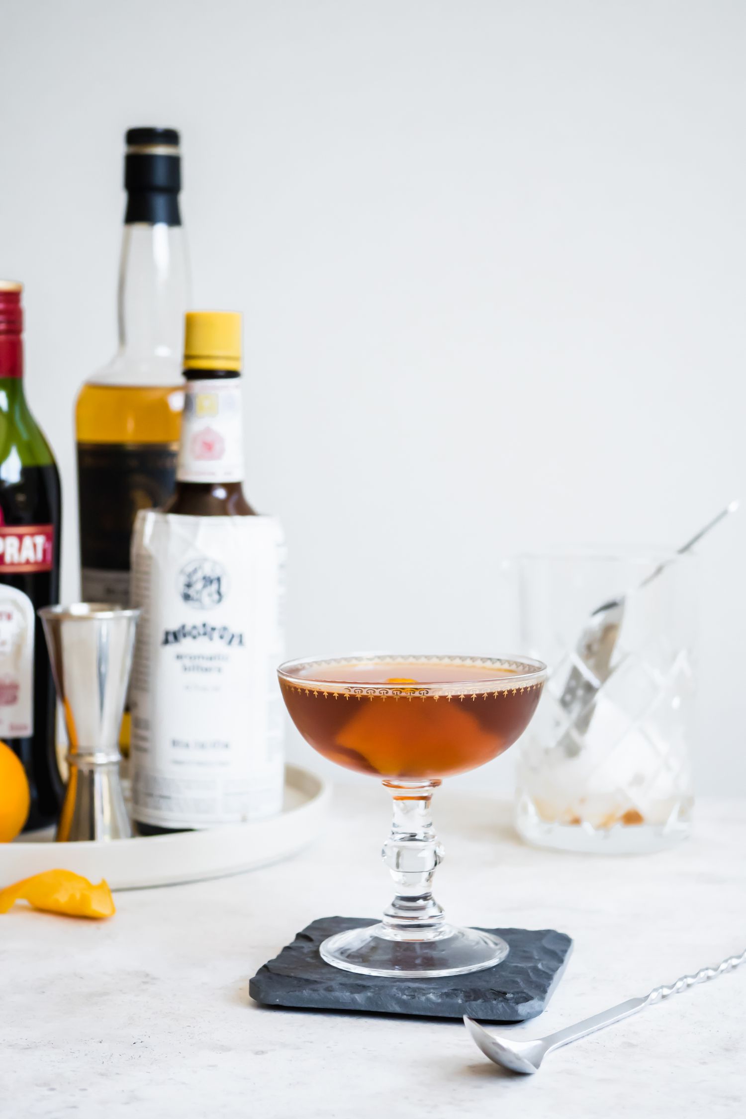 Rob Roy in Cocktail Glass on Top of Granite Coaster Surrounded by a Cocktail Mixing Glass and and a Tray with Recipe Ingredients (Vermouth, Whiskey, and Orange)