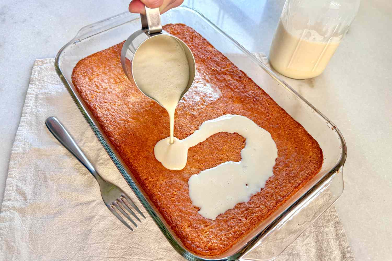 Milk being poured from a measuring cup onto a baked cake in a glass dish, with a fork and jar nearby