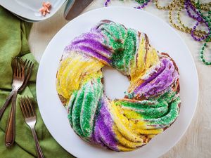 Top view of a homemade king cake on a white plate. Forks and a green linen are to the left. A small plastic baby figurine is above as well as Mardi Gras beads