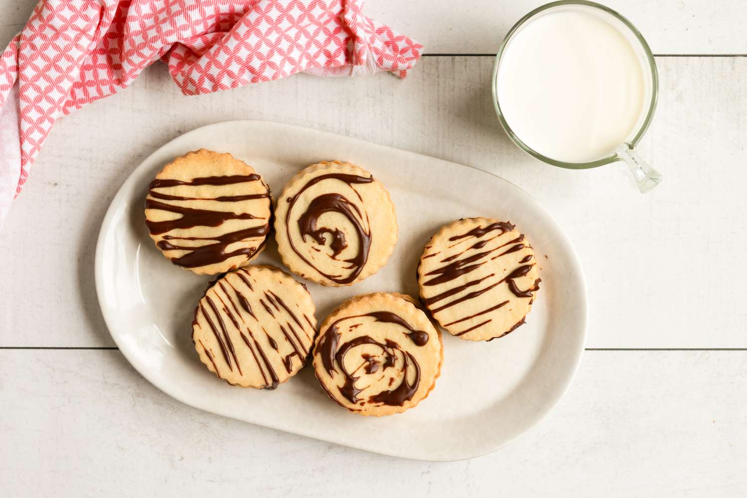Plate of cookies with chocolate drizzle and a glass of milk on a white table with a patterned cloth nearby