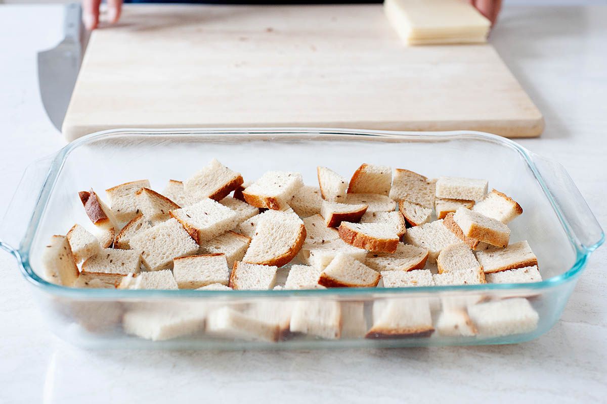 Cubed rye bread is in a glass casserole dish and behind it is a blonde cutting board with a stack of swiss cheese.