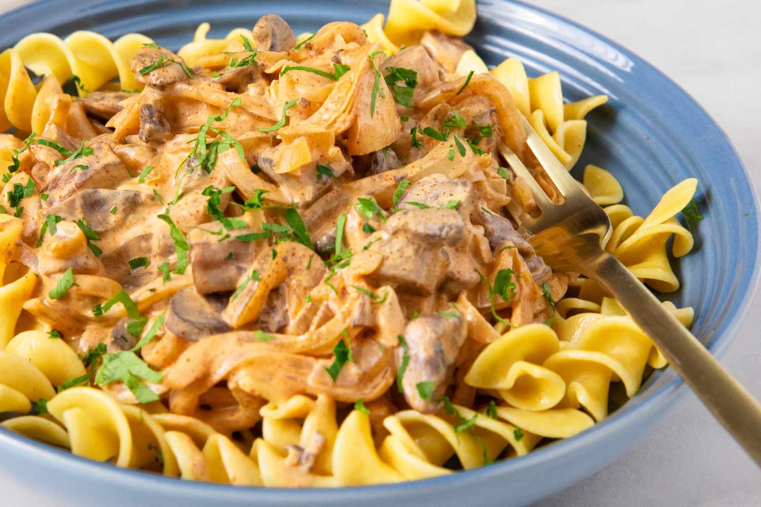 A plate of mushroom stroganoff served over egg noodles garnished with chopped parsley and accompanied by a fork