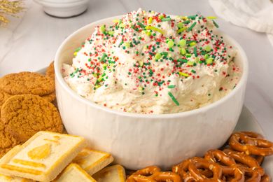 A bowl of festive dessert dip topped with sprinkles surrounded by cookies and pretzels on a serving tray