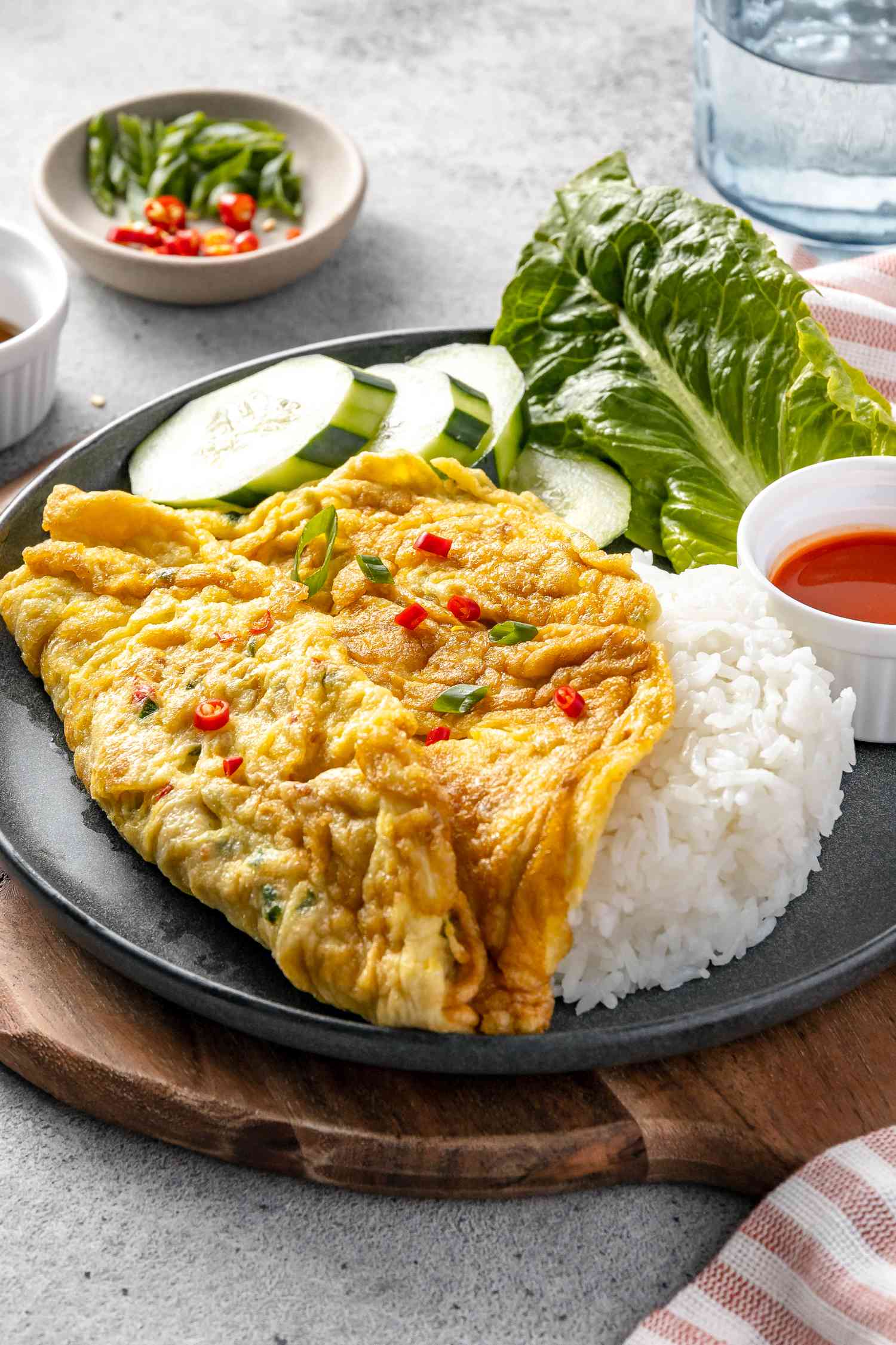 Plate of Kai Jeow (Thai Omelet) with Cucumber Slices, Romaine Leaves, Rice, and a Small Bowl of Sriracha on a Wooden Circular Serving Tray and Next to a Kitchen Towel, a Glass, and a Small Bowl with Toppings