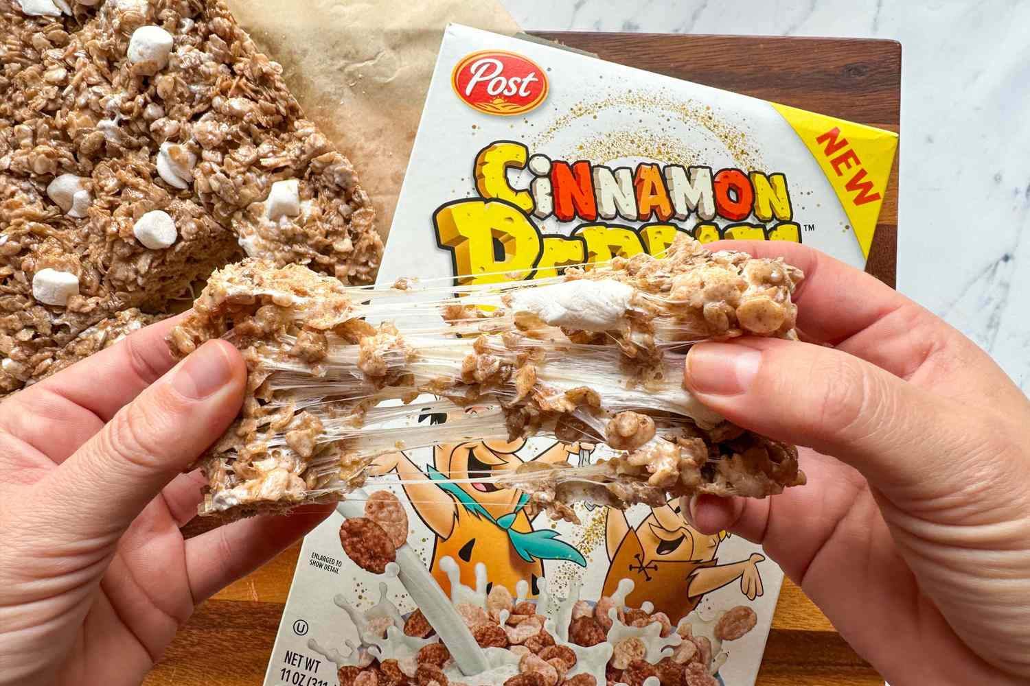 Hands pulling apart a gooey cinnamon flavored cereal treat in front of its box on a wooden surface