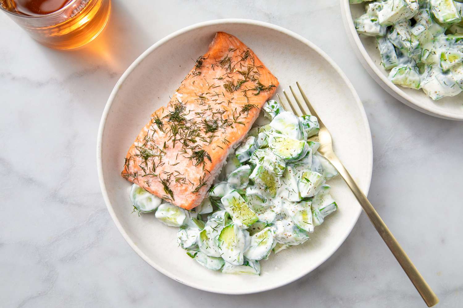 A plate with a piece of cooked salmon and a cucumber salad with a creamy dressing, accompanied by a fork