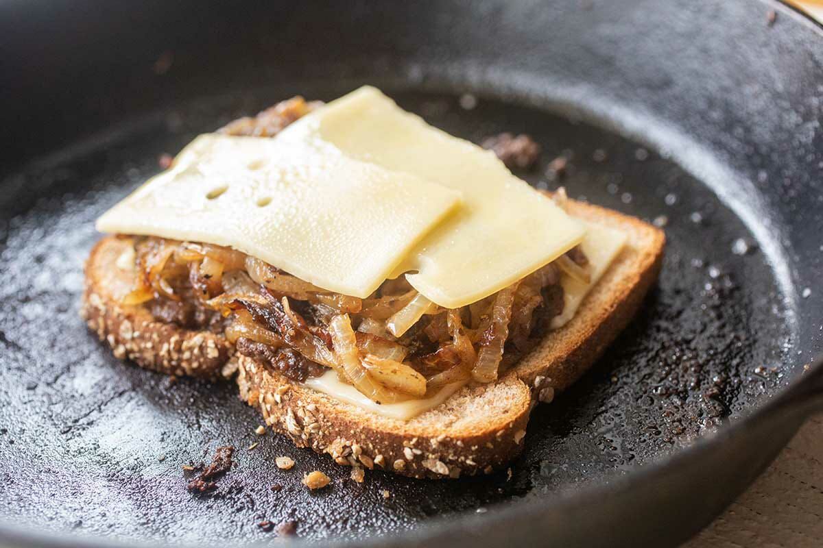 Perfect patty melt being assembled in a cast iron skillet. The bottom slice of bread is topped with cheese, caramelized onions and another slice of cheese.