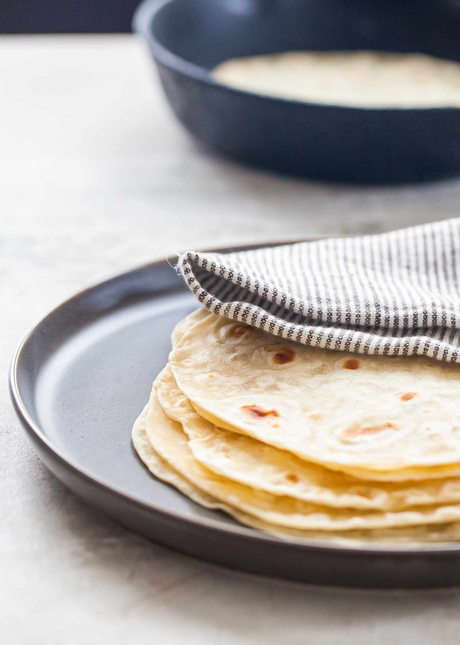 A stack of homemade flour tortillas on a platter covered by a striped towel. A cast iron skillet with another tortilla inside is in the background.