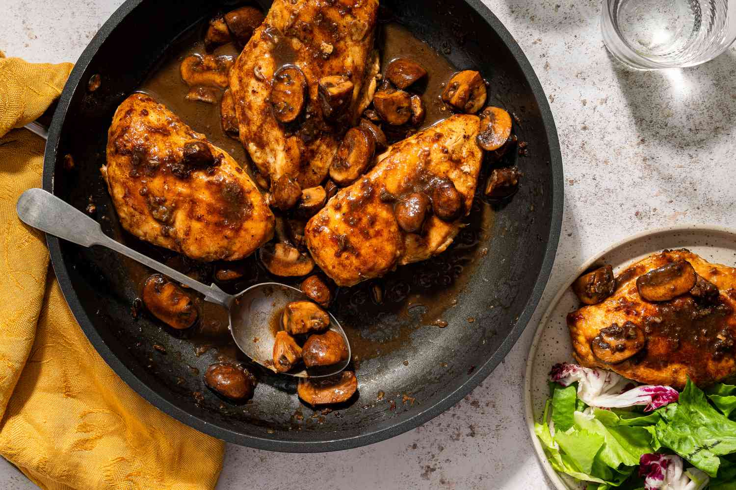 Three chicken breasts with balsamic mushrooms in a frying pan, with one chicken breast put on a plate with greens next to the pan