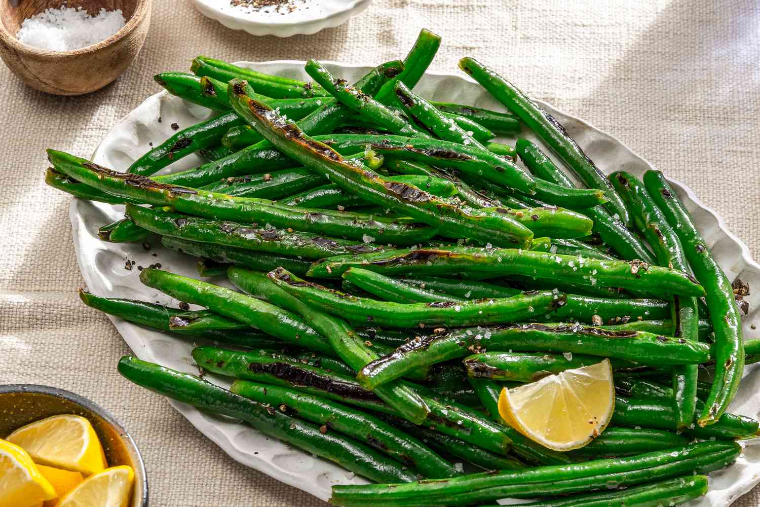 Overhead shot of a white oval serving plate with slightly charred pan fried green beans, sprinkled with coarse sea salt and garnished with a lemon wedge