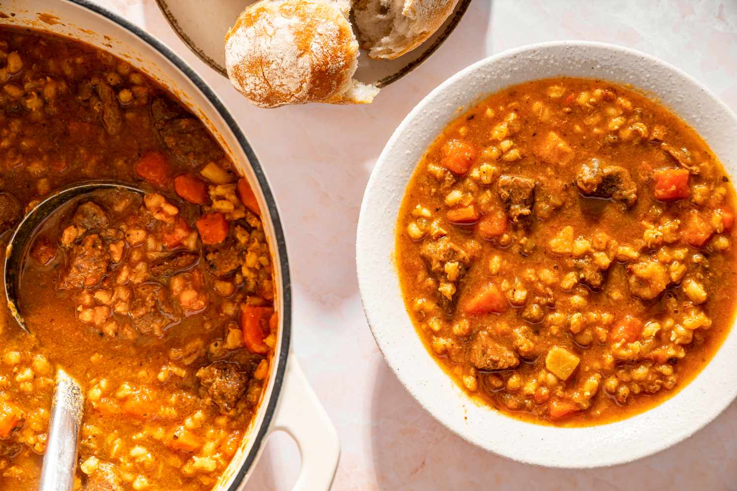 Beef barley soup served in a pot with a ladle and in a white bowl bread roll beside it