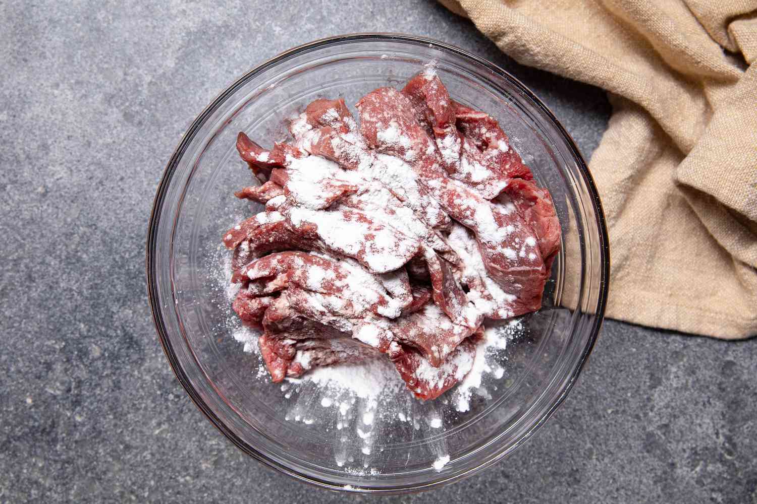 Baking Soda Added to Beef Slices in a Bowl Next to a Pale Beige Kitchen Towel