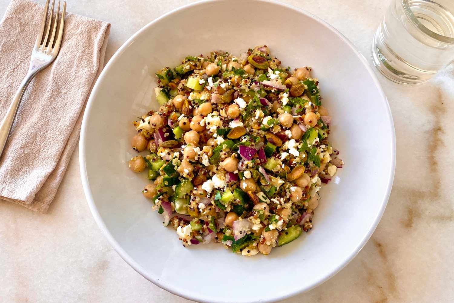Overhead photo of the perfect salad in a wide bowl on a marble counter, showing quinoa, chickpeas, cucumber, red onion, and feta