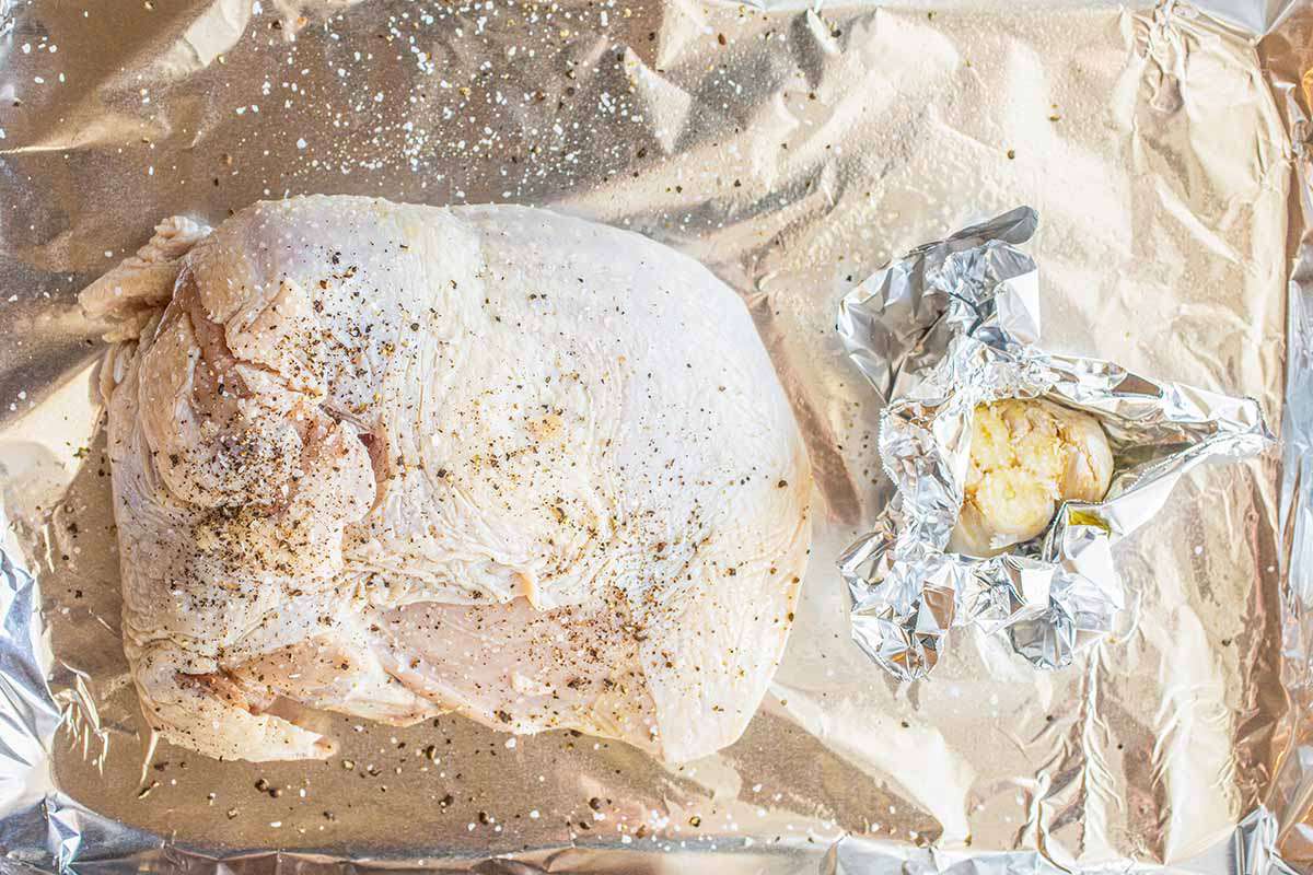 Turkey breast coated with salt and pepper on a foil lined baking tray. A head of garlic is wrapped in foil next to the turkey.
