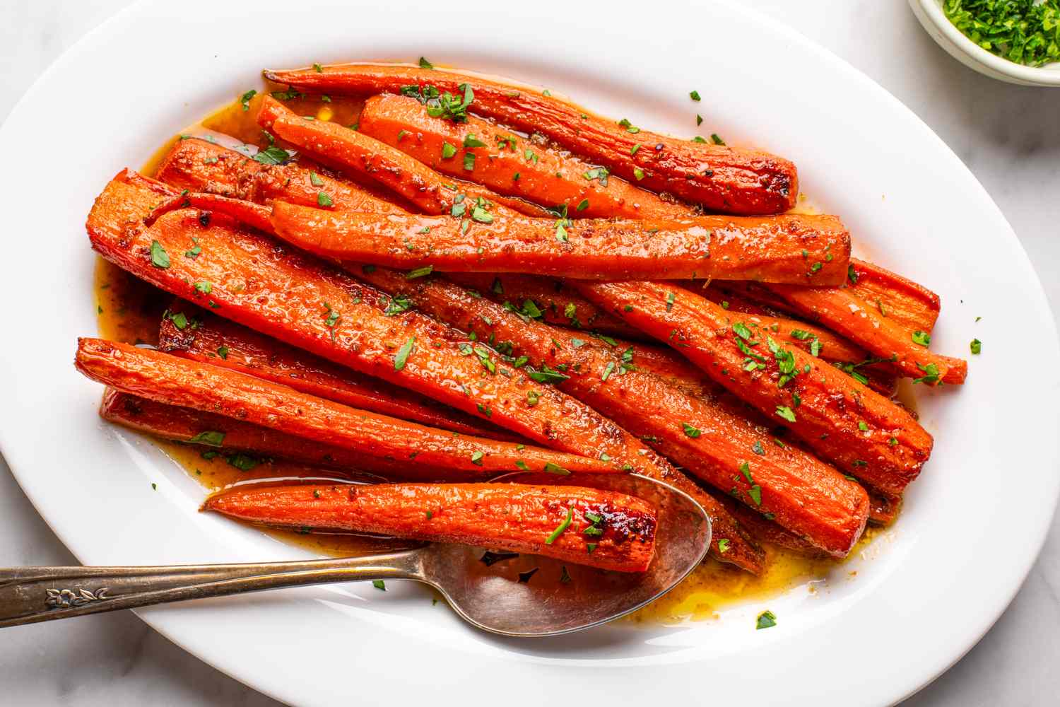Dish of cooked carrots on a white plate with chopped herbs and a serving spoon