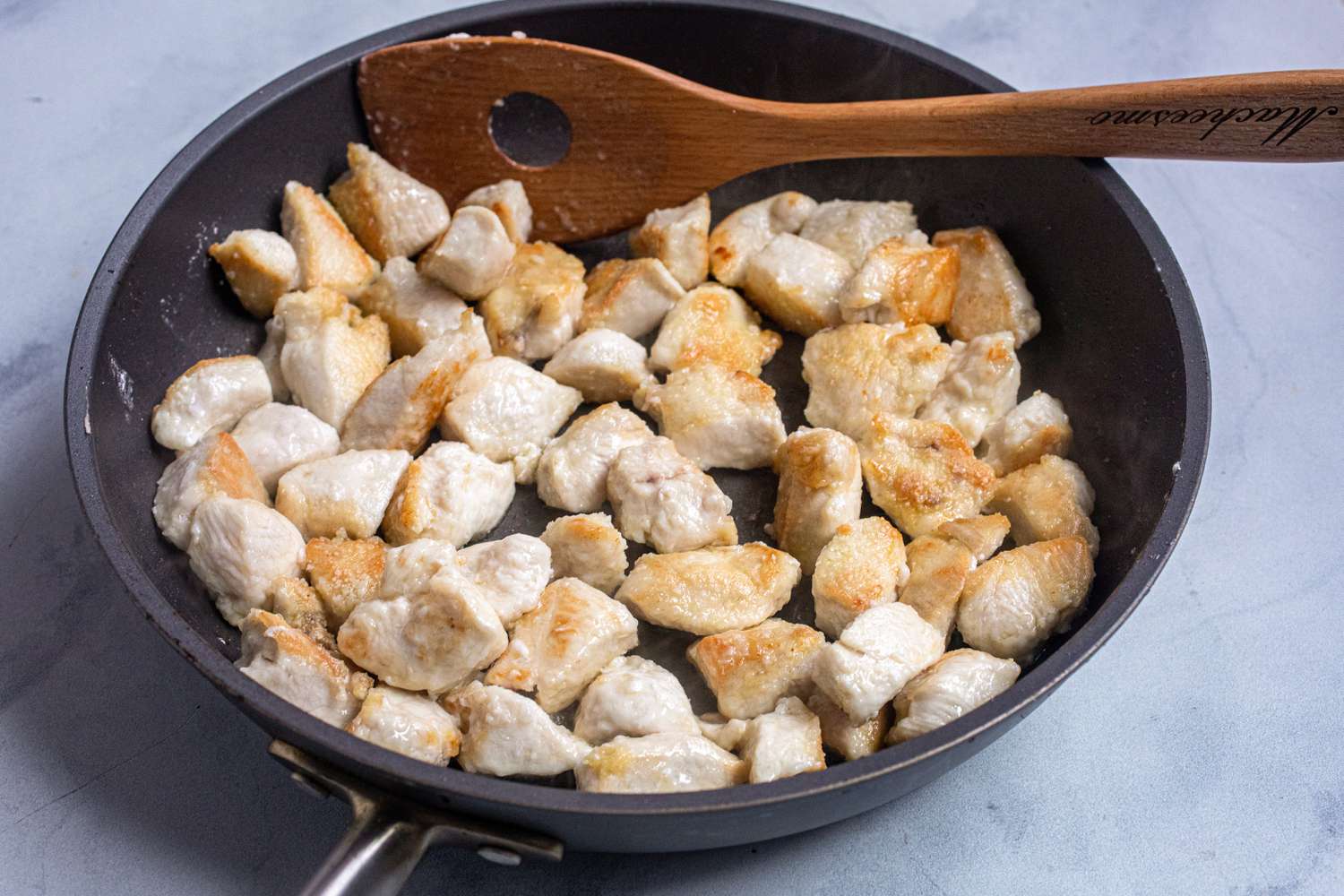 A skillet with chicken browning and a wooden spoon to make Sweet and Tangy Bourbon Chicken.