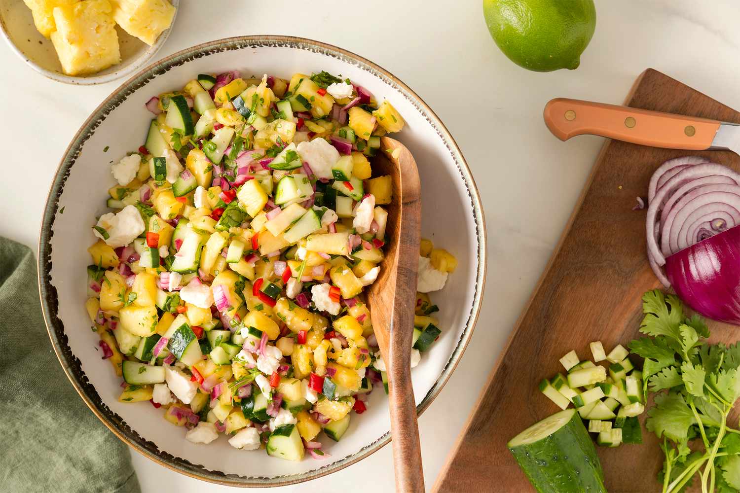 Pineapple Cucumber Salad with Feta in a serving bowl with a wooden spoon, cutting board with finely chopped cucumber, sliced red onion, cilantro, lime, bowl of fresh pineapple chunks
