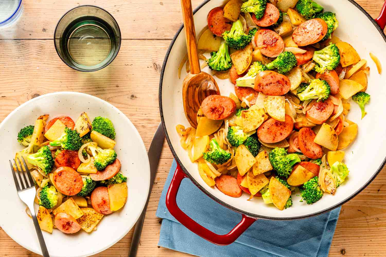 Overhead view of a white bowl of Kielbasa, Potato, and Broccoli Skillet with a fork next to a skillet on a blue fabric napkin and a drinking glass 
