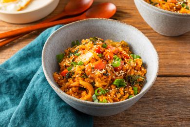 Bowl of kimchi and spam fried rice at a table setting with another serving in a bowl, a table linen, two spoons, and a plate with a fried egg