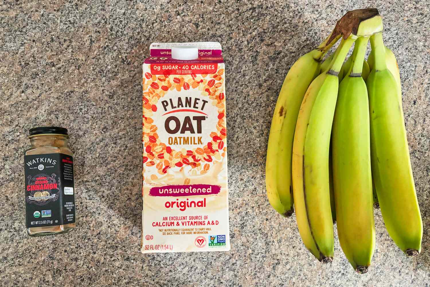 A bundle of bananas next to a carton of oat milk and a jar of cinnamon on a countertop