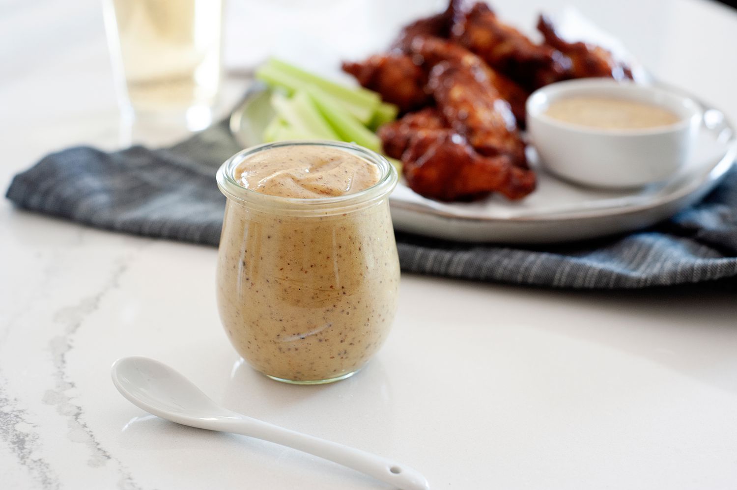 Side view of homemade honey mustard in a glass jar with a plate of chicken wings behind it.