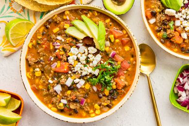 Stovetop Taco Soup in a Bowl Topped with Avocado Slices, Cotija Cheese, and Cilantro and Surrounded by Bowls of Toppings