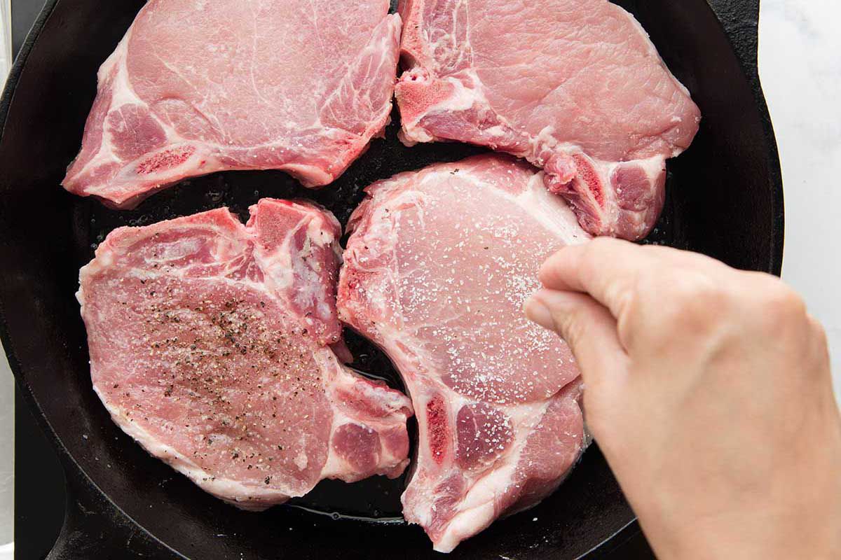 Pork chops in a cast iron skillet with a hand sprinkling salt and pepper over the chops.