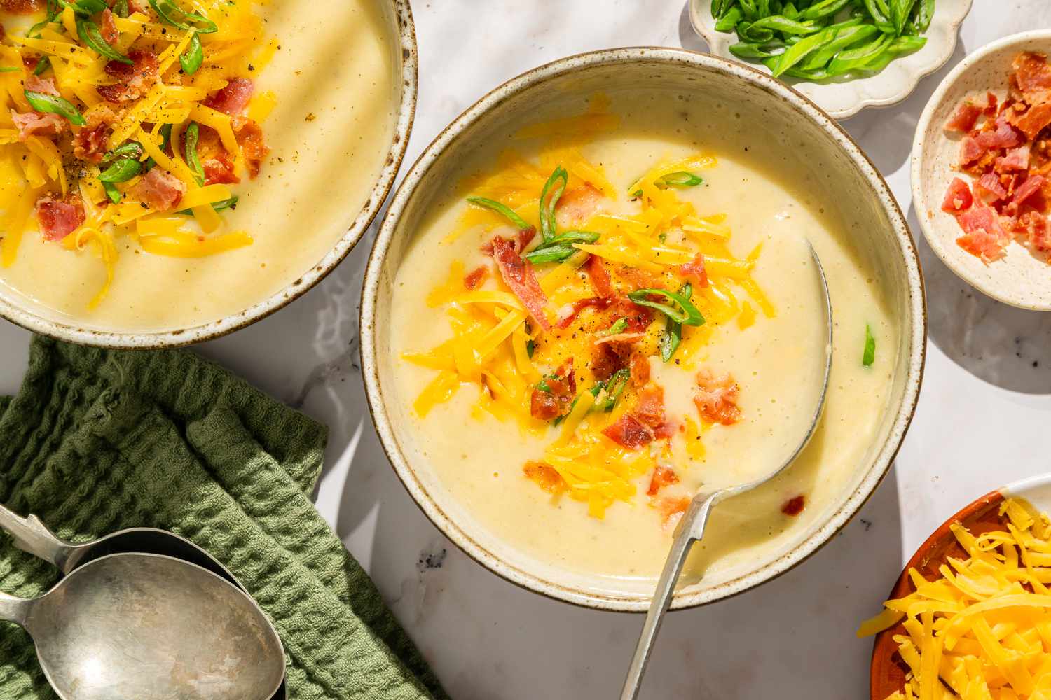 Overhead view of two bowls of potato soup topped with bacon, shredded cheese and green onions next to a bowls of bacon, green onions and shredded cheese