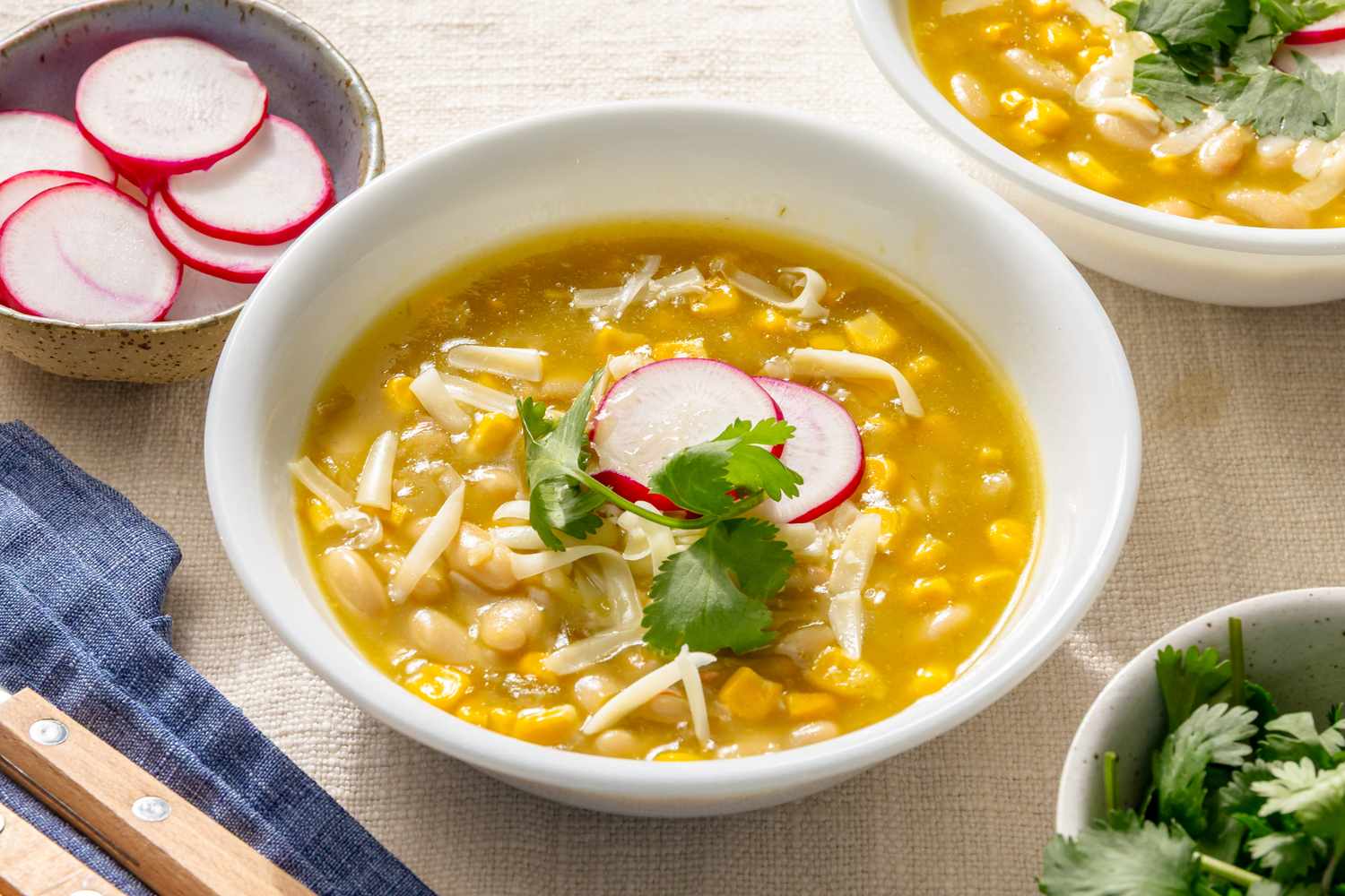 A bowl of chili verde garnished with radish slices and cilantro surrounded by additional toppings in small dishes