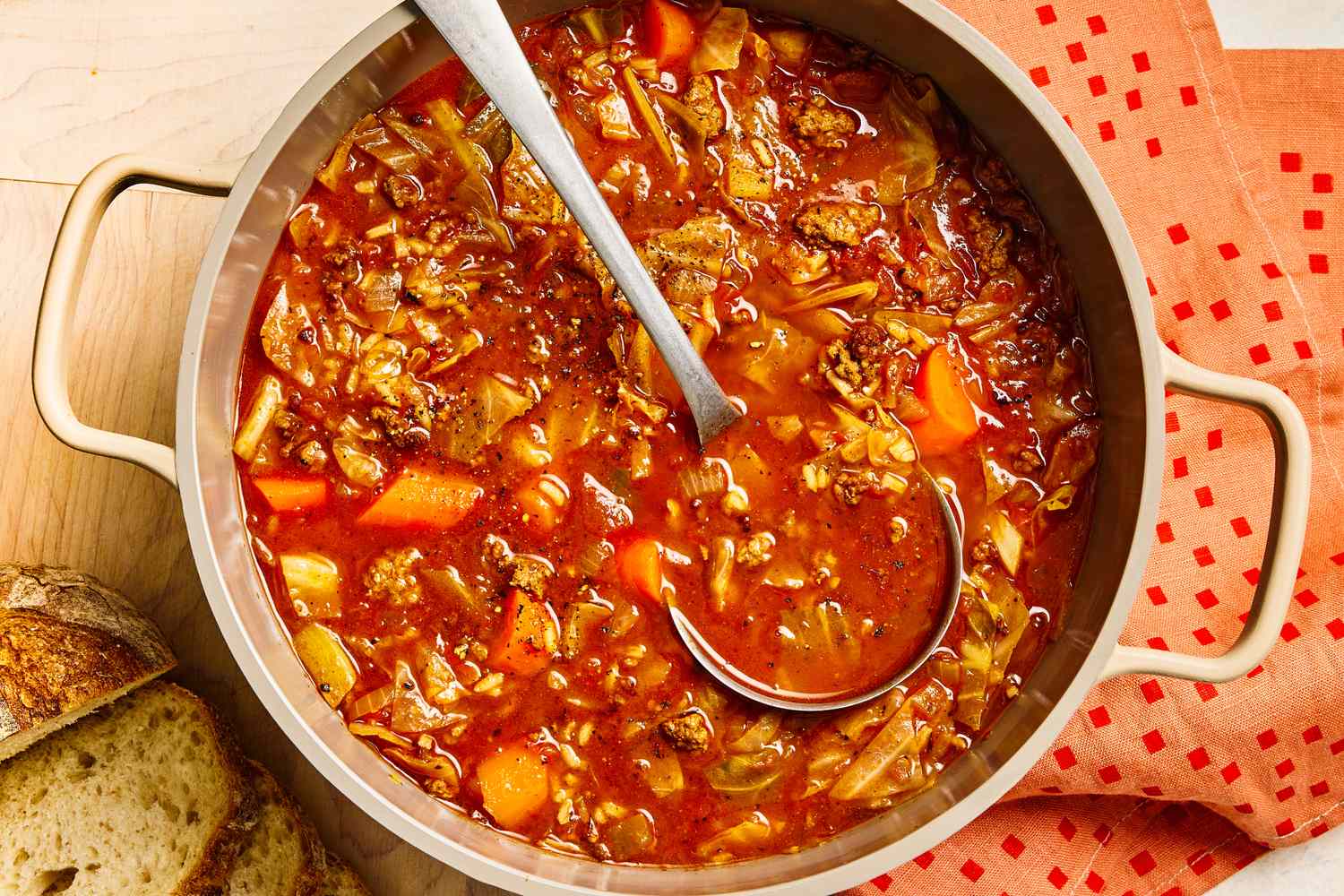 Pot of cabbage roll soup with a ladle on a wooden table accompanied by bread